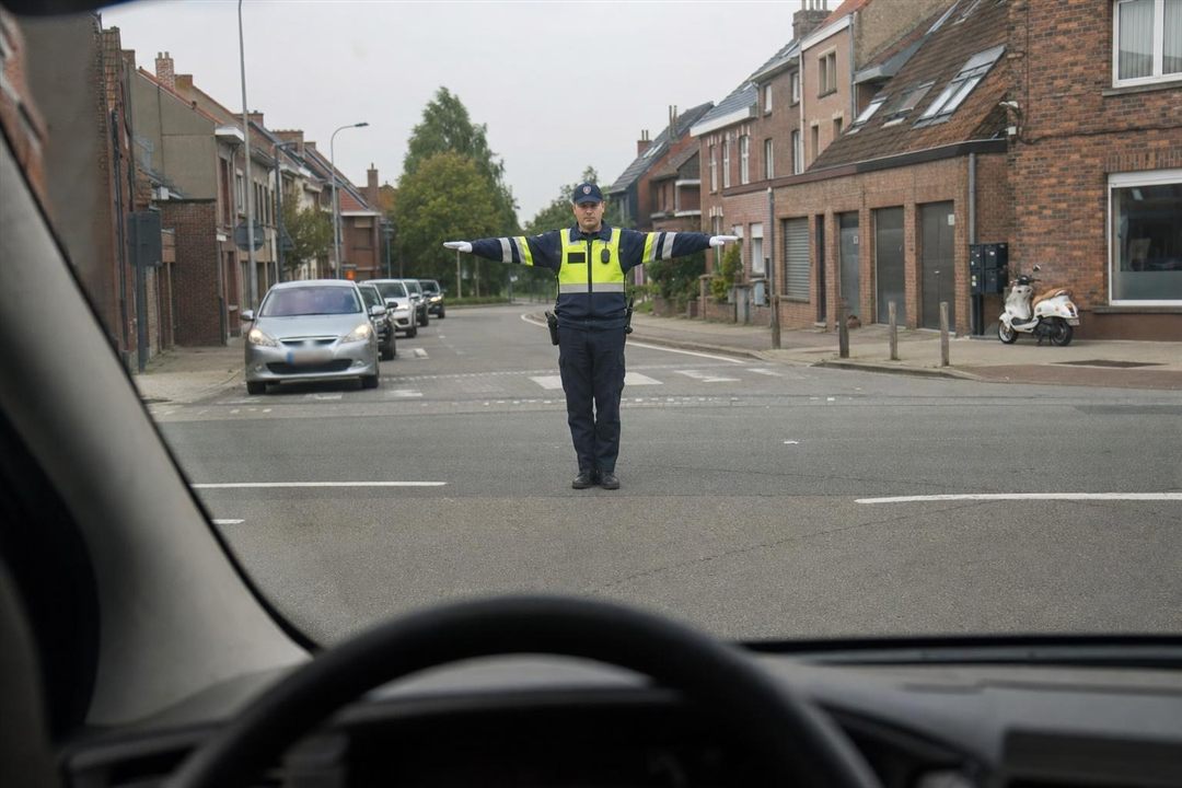 Agent belge au centre du carrefour, bras tendus à l'horizontale, vu à travers le pare-brise